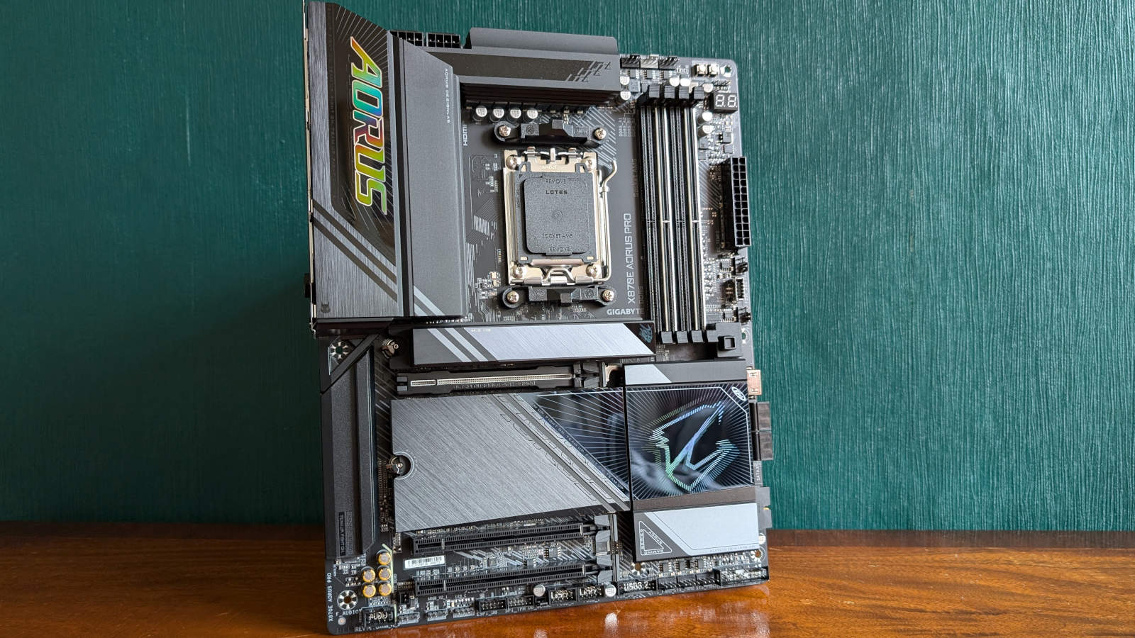 Wide shot of X870E Aorus Pro, standing upright on a wooden surface against a dark green backdrop.