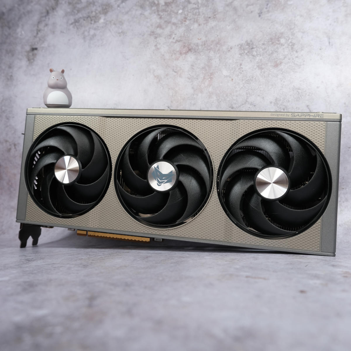 Sapphire Nitro+ Radeon RX 9060 XT 16GB sits vertically against a pearlescent background with a small mouse figurine (top left) standing on it.