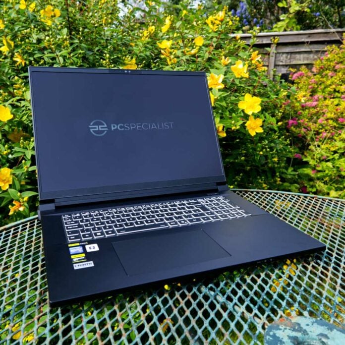 PCSpecialist Recoil 18 gaming laptop on a garden table against a backdrop of flowers.