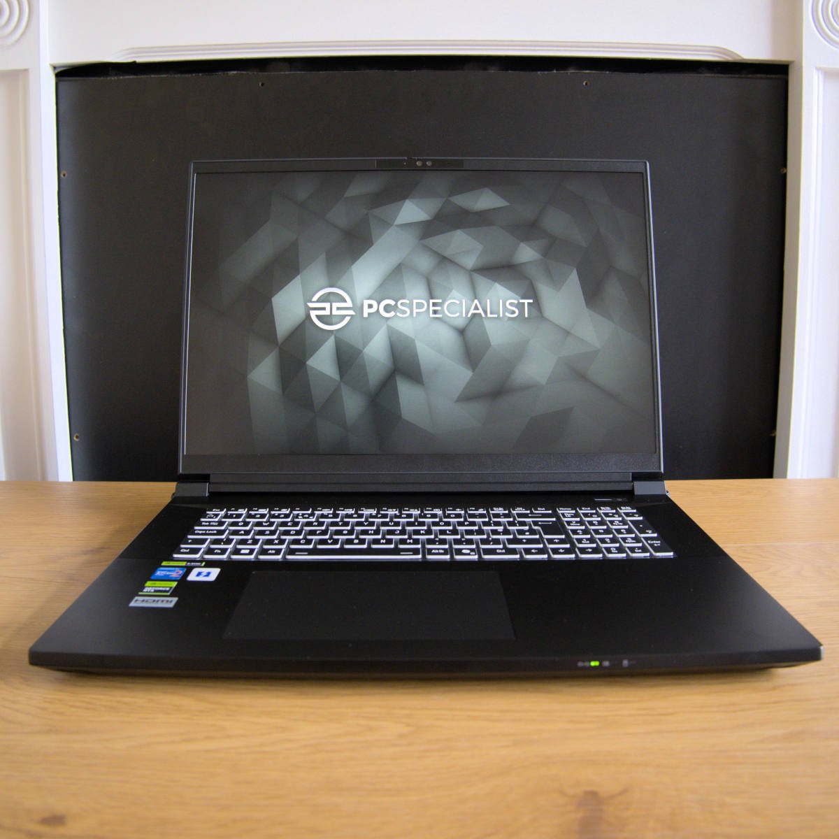 PCSpecialist Recoil 18 laptop, sat atop a wooden surface with a fireplace behind it.
