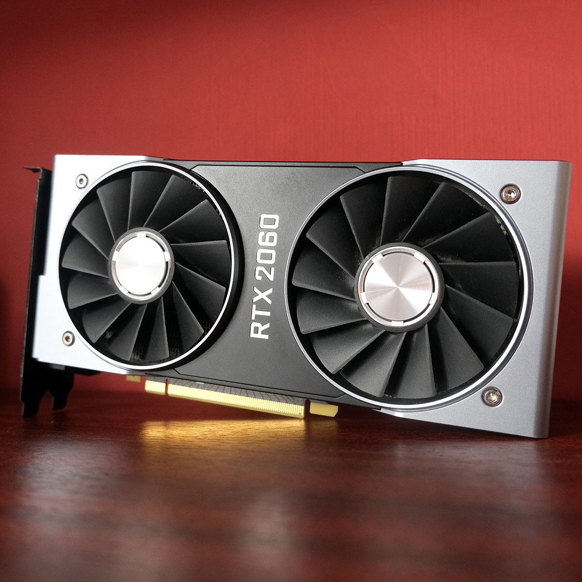 Nvidia GeForce RTX 2060 Founders Edition graphics card standing upright on a dark wood surface, against a red backdrop.
