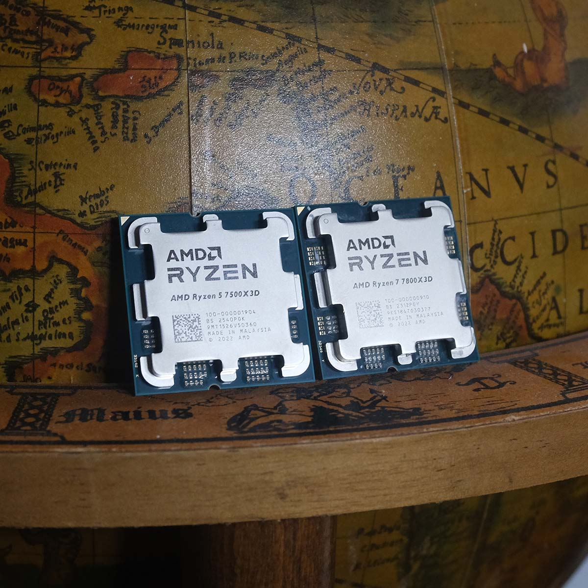 An AMD Ryzen 5 7500X3D (left) standing upright next to a Ryzen 7 7800X3D (right) against a wooden globe.