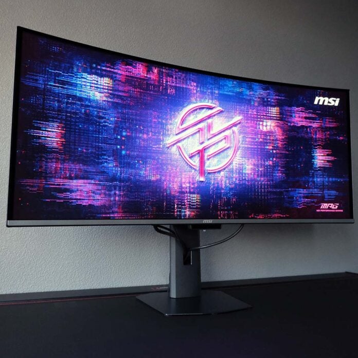 The MSI MPG 341CQR QD-OLED X36 standing atop a desk, displaying an MPG desktop wallpaper.