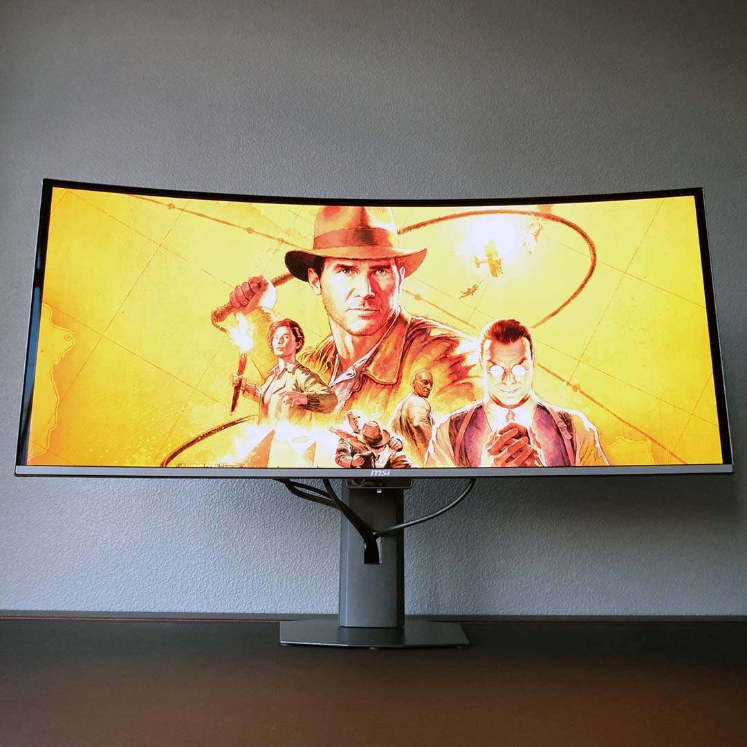 The MSI MPG 341CQR QD-OLED X36 standing atop a desk, displaying an Indiana Jones wallpaper.