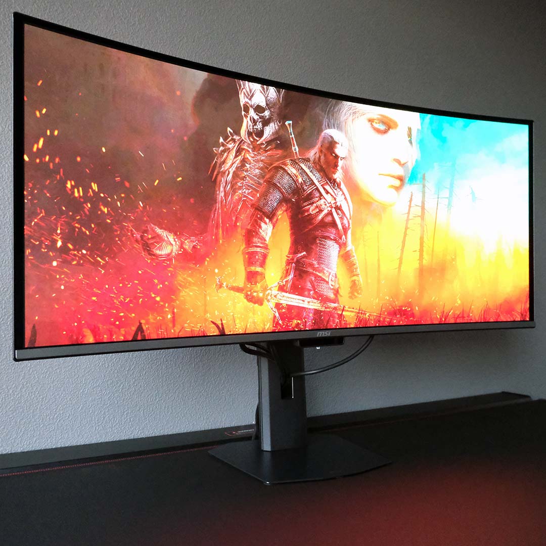 The MSI MPG 341CQR QD-OLED X36 standing atop a desk, displaying a Witcher 3 wallpaper.